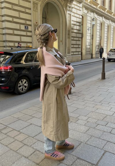 Trench de color beige sobre un suéter rosa claro colocado sobre los hombros. Llevado con jeans azules y zapatillas de plataforma rosas con rayas marinas.