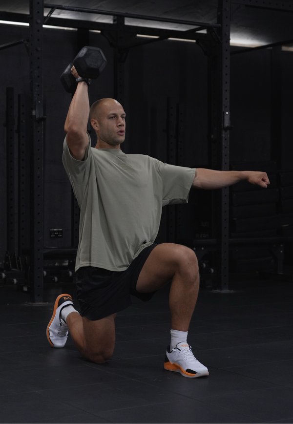 Homme à genoux sur un genou dans une salle de sport, levant un haltère au-dessus de la tête avec un bras tout en tendant l'autre bras vers l'avant pour l'équilibre.