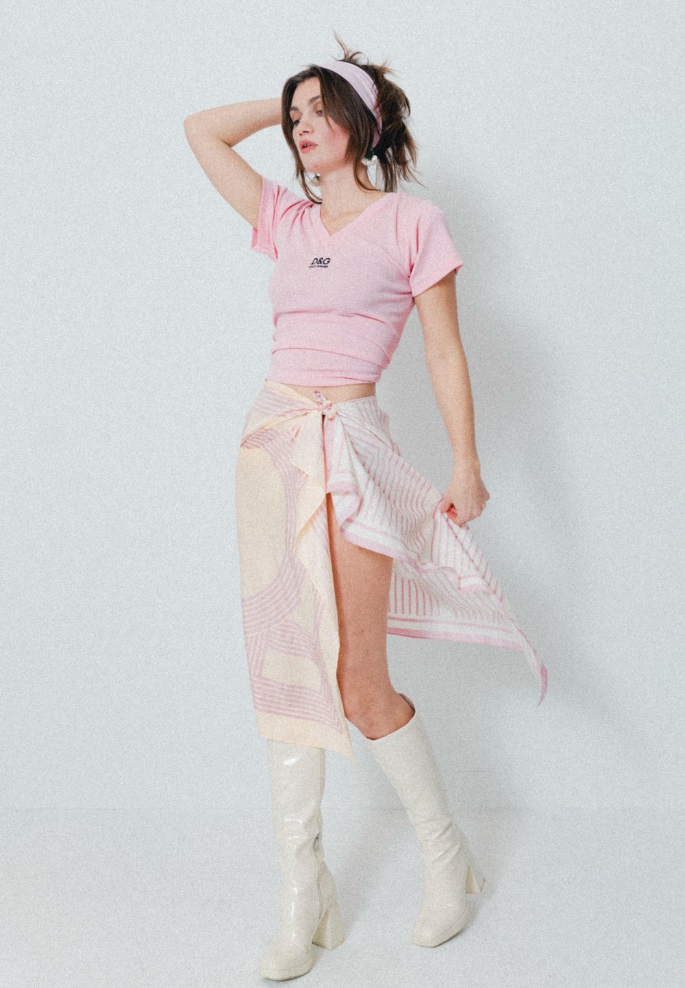 T-shirt rose ajusté avec un petit logo, associé à une jupe portefeuille à motifs rose clair et crème et des bottes blanches laquées jusqu'au genou.