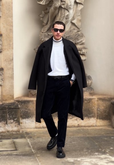 Abrigo negro llevado sobre un suéter de cuello alto blanco, combinado con pantalones de pana azul marino y zapatos negros. Los accesorios incluyen gafas de sol negras y un reloj.