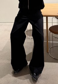 Personne portant un pantalon large noir et des chaussures noires à bouts pointus, debout près d'une table en bois avec des chaises brunes sur un sol en béton.