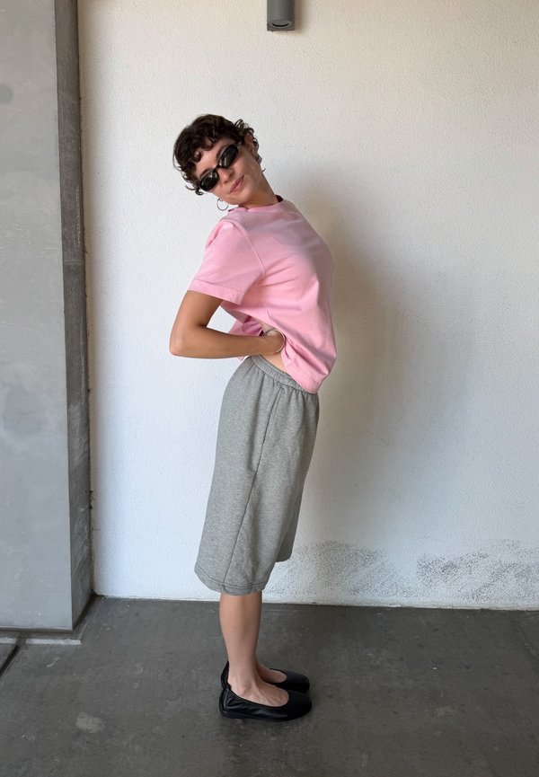 Persoon die een roze t-shirt, grijze kniebroek en zwarte platte schoenen draagt, staat zijwaarts met handen op de onderrug tegen een witte muur.