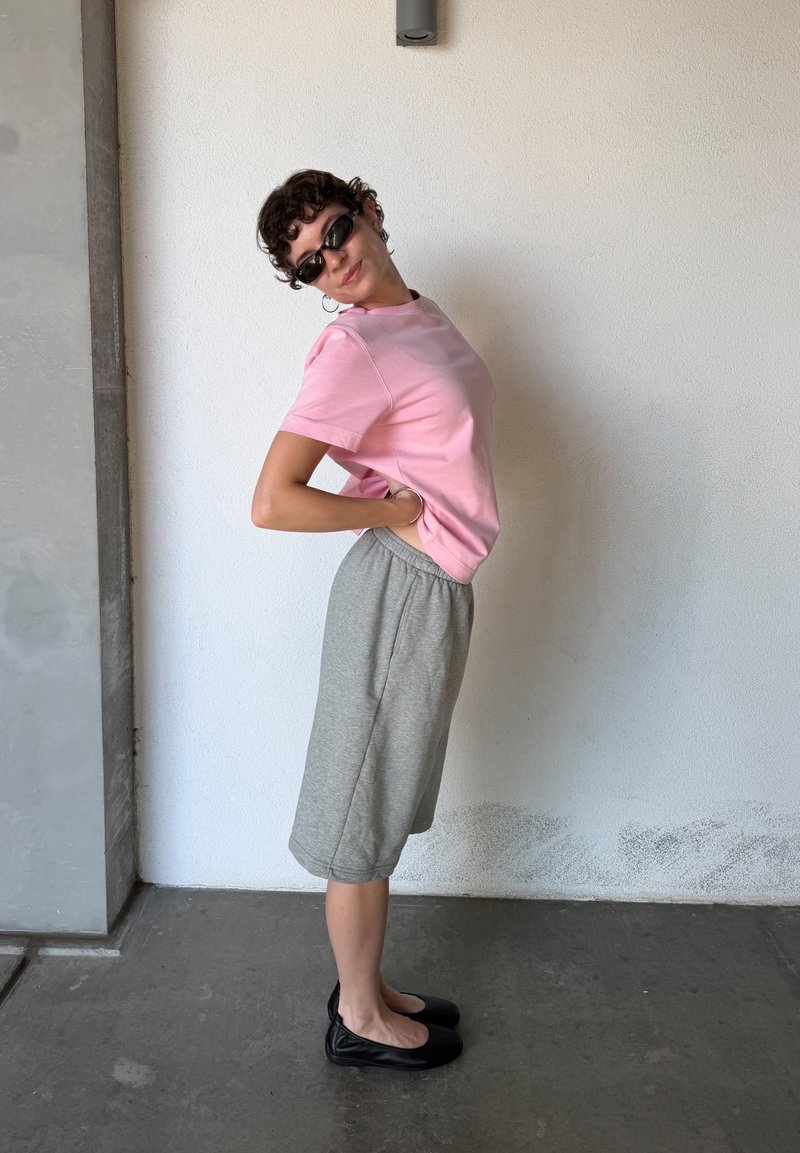 Personne portant un t-shirt rose, un short gris jusqu'aux genoux et des ballerines noires, debout de profil les mains sur le bas du dos contre un mur blanc.