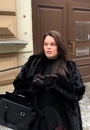 Mujer con abrigo de piel negro y guantes sosteniendo gafas de sol, sentada junto a una puerta marrón con un gran bolso negro a su lado al aire libre.