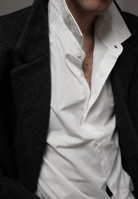 Homme portant une chemise blanche déboutonnée et un manteau en laine gris foncé, montrant des poils de poitrine sur un fond neutre.