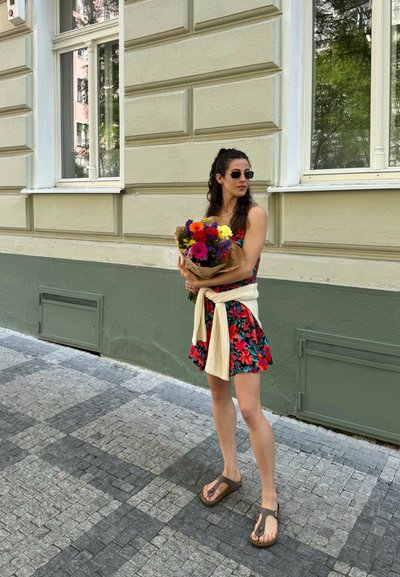 Mujer con un vestido floral de patrones rojos y verdes, sosteniendo un ramo de flores coloridas envuelto en papel marrón, usando gafas de sol y sandalias.