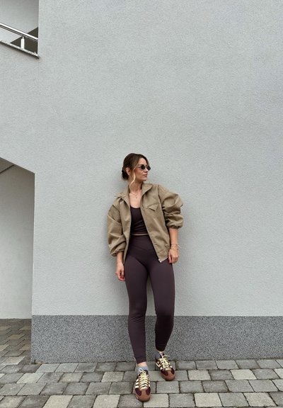Chaqueta tipo plumas beige, leggings ajustados marrones y zapatillas marrones con cordones amarillos. La superficie es una pared gris y el suelo está pavimentado con piedras.