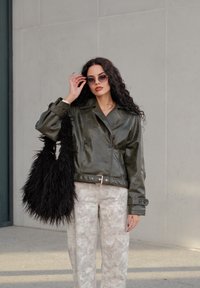 Olivově zelená krátká bunda z imitace kůže s dvouřadým zapínáním, spárovaná se světle šedými vzorovanými širokými kalhotami a černou chlupatou kabelkou.