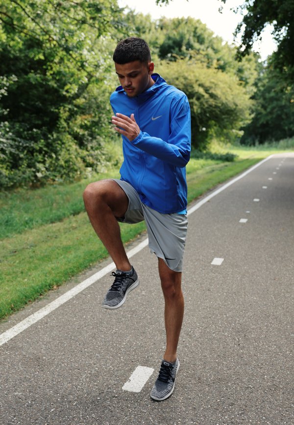 Blauem Nike-Jacke, grauen Shorts und schwarzen Sportschuhen. Die Person hebt ein Knie, während sie auf einem gepflasterten Weg joggt, umgeben von Grünflächen.