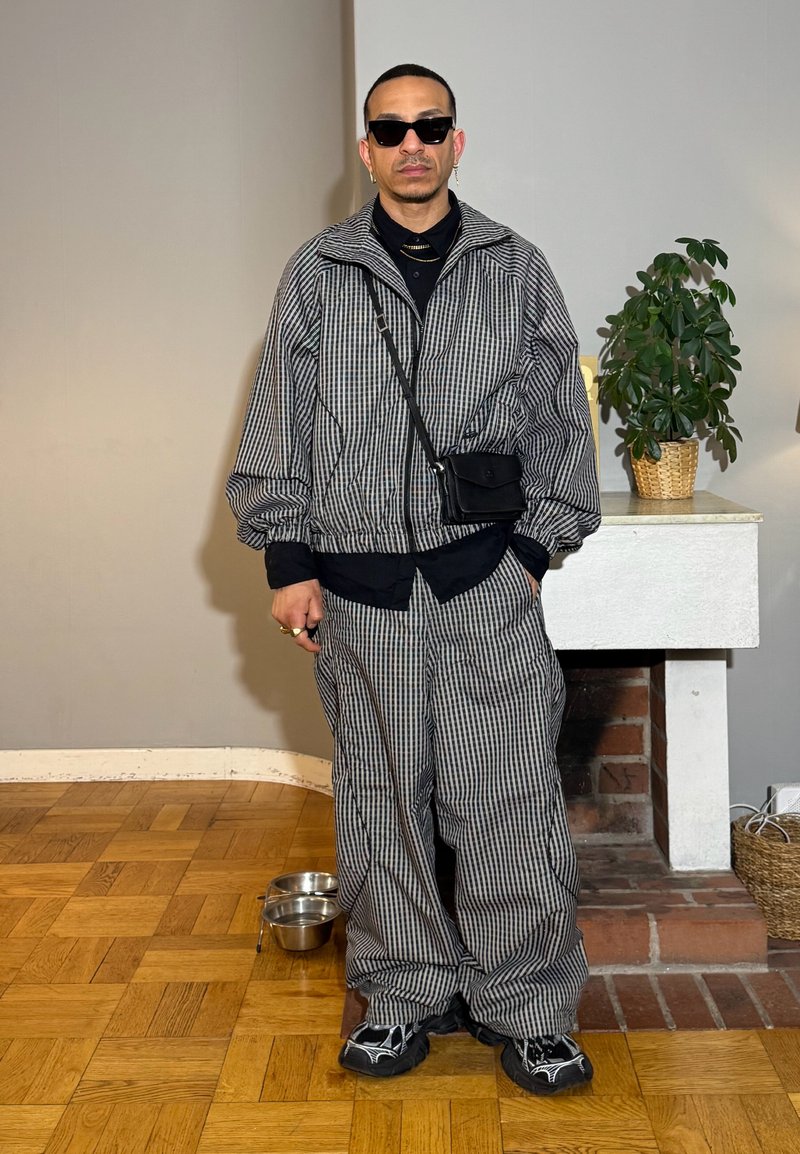 Homme portant une veste et un pantalon à carreaux noir et blanc, lunettes de soleil noires, debout à l'intérieur sur un sol en bois près d'une cheminée avec une plante.
