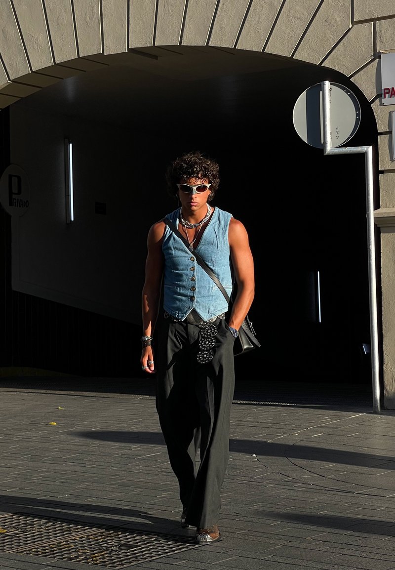 Τζην γιλέκο με κουμπιά, σκούρες φαρδιές παντέλες, μεταλλικά αξεσουάρ και λευκά γυαλιά ηλίου. Στεκόμενος σε έναν φωτισμένο διάδρομο με ελάχιστες λεπτομέρειες.