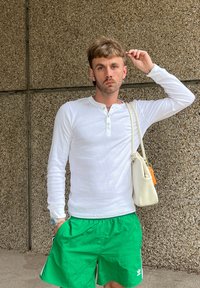 Weißes Langarm-Henley-Shirt, grüne Shorts und eine weiße Handtasche mit kurzem Tragriemen. Der strukturierte Hintergrund ist grau und grob.