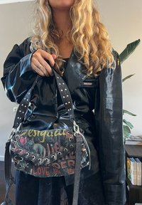 Femme aux longs cheveux blonds bouclés portant une veste noire brillante et tenant un sac à main style graffiti avec des mots et des images, debout à l'intérieur.