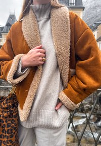 Άτομο που φοράει μπεζ μπουφάν με εσωτερική επένδυση shearling πάνω από γκρι πλεκτό πουλόβερ, κρατώντας τσάντα με λεοπάρ σχέδιο, στέκεται κοντά σε μια κουπαστή με πέτρινα κτίρια στο φόντο.