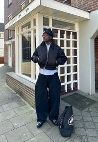 Schwarze Bomberjacke mit silbernem Reißverschluss, getragen über einem hellblauen gestreifen Hemd. Weite schwarze Hose und schwarze Schuhe. Schwarze Tragetasche auf dem Boden.