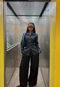 Schwarze Jeansjacke mit hohem Kragen, kombiniert mit weiten schwarzen Hosen. Der Hintergrund zeigt metallische Aufzugwände und gelbe Akzente.