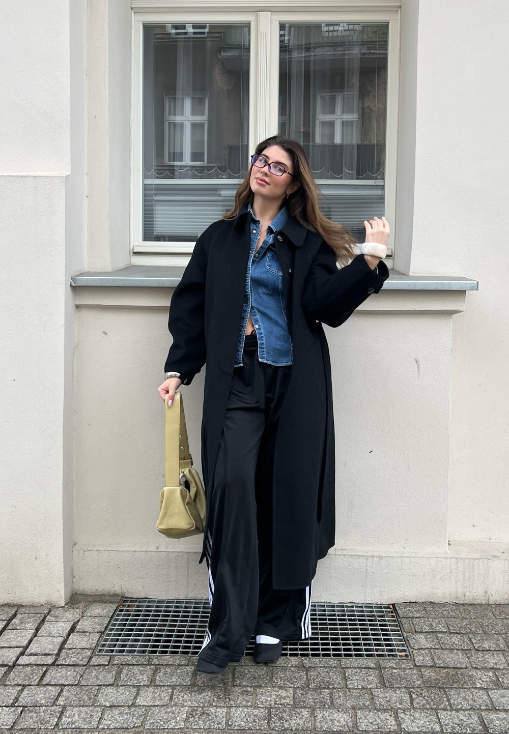 Long black coat over a denim shirt, black wide trousers, and sporty shoes. Holding a soft, light green bag. Standing near a window.