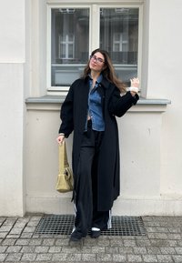 Long black coat over a denim shirt, black wide trousers, and sporty shoes. Holding a soft, light green bag. Standing near a window.