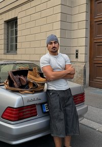 Braune Schnürstiefel, eine braune Leder Tasche und eine graue Mütze liegen auf einem silbernen Auto. Der Mann trägt ein graues T-Shirt und dunkle Shorts.