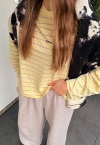 Άτομο με κίτρινο ριγέ μακρυμάνικο πουκάμισο, ανοιχτομπεζ παντελόνι φόρμας και μα ύρο-άσπρο φλις μπουφάν περασμένο πάνω από τον έναν ώμο.