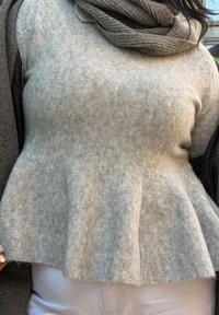 Pull en maille grise avec un design péplum évasé, présentant un tissu doux et texturé ainsi qu'une écharpe douillette pliée dans un gris plus foncé.