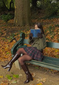 Strikket olivengrønn genser, rutet skjørt i marineblått og rødt, brune knelange støvler, holder en blå bok, sitter på en grønn benk blant høstløv.