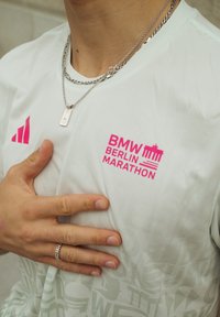Cămașă sport albă cu textură din plasă, având logo-ul roz "BMW BERLIN MARATHON" și trei dungi roz. Se văd lanțuri și un inel argintii.