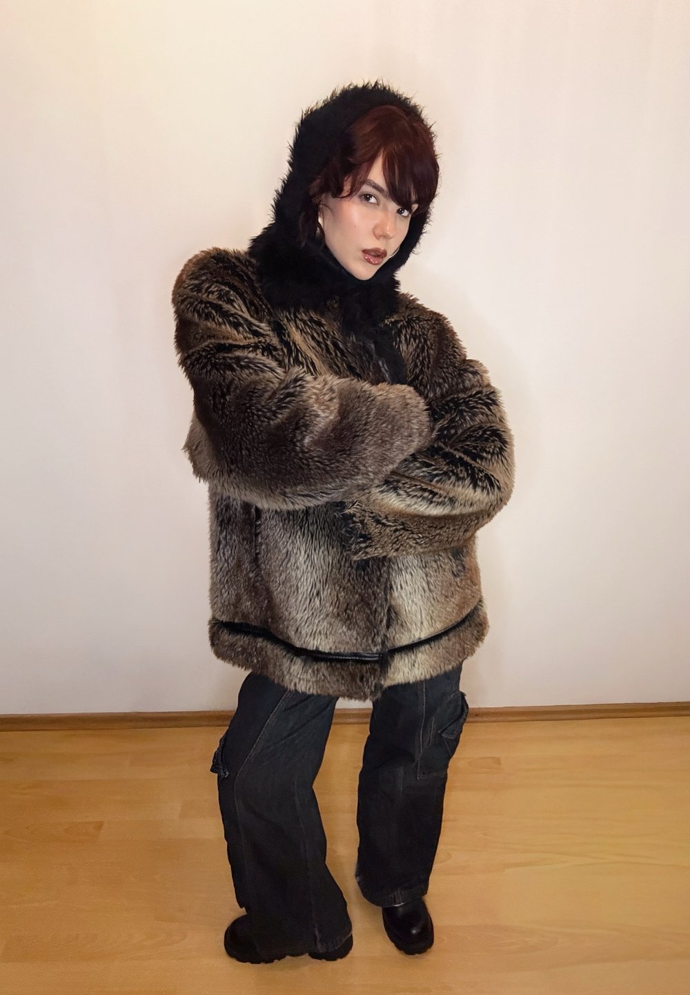 Manteau en fausse fourrure marron avec capuche noire, coupe oversize, présentant une texture côtelée et un cordon de serrage. Associé à un pantalon cargo noir ample et des bottes.