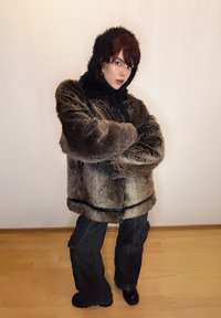 Manteau en fausse fourrure marron avec capuche noire, coupe oversize, présentant une texture côtelée et un cordon de serrage. Associé à un pantalon cargo noir ample et des bottes.