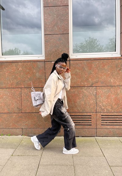 Persona que lleva puesto un chaleco beige, una camisa blanca y pantalones cargo negros con desgastes. Mochila gris con un hebilla. Gafas transparentes y zapatillas blancas.