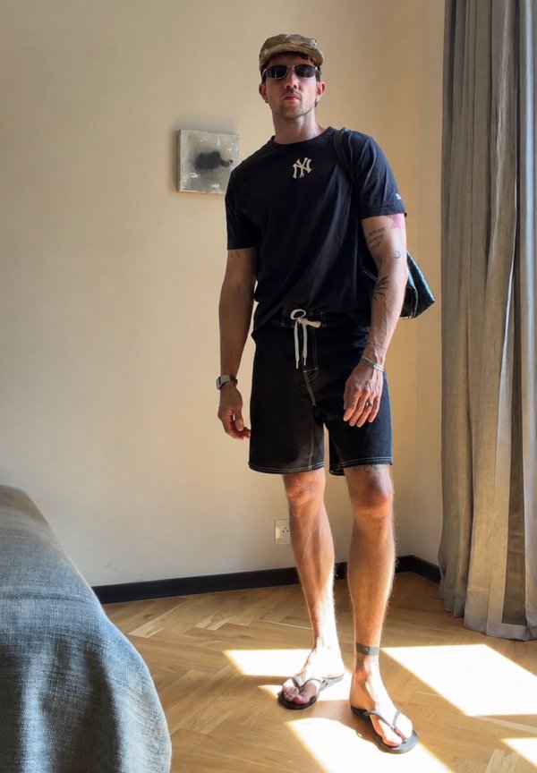 Man wearing black New York Yankees shirt, black shorts, flip-flops, cap, sunglasses, and carrying a backpack standing in a sunlit room.