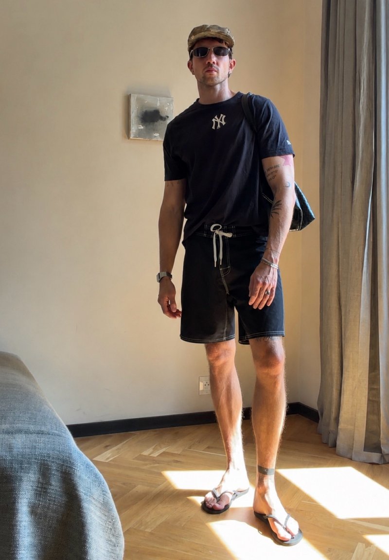 Homme portant un t-shirt noir des New York Yankees, un short noir, des tongs, une casquette, des lunettes de soleil et un sac à dos, debout dans une pièce ensoleillée.