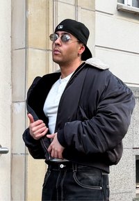 Fekete bombázódzseki gyapjúbélelt gallérral, alatta fehér ing, valamint sötét farmernadrág. Kiegészítve napszemüveggel és fekete sapkával.