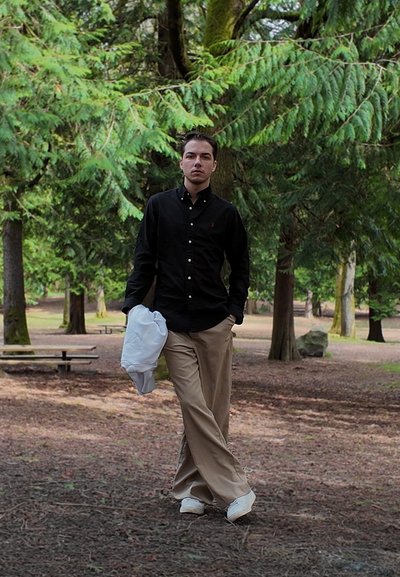 Joven caminando en un parque forestal, vestido con camisa negra, pantalones beige, zapatos blancos, sosteniendo una chaqueta blanca en una mano.