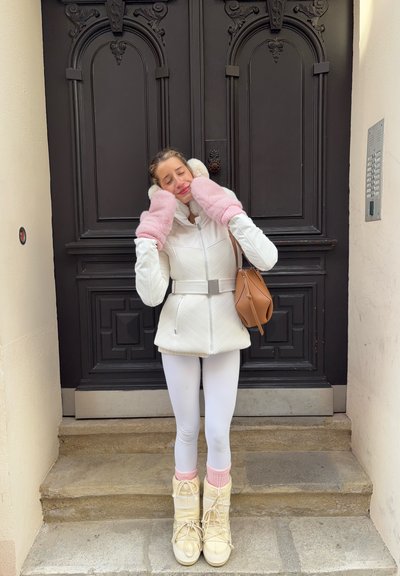 Chaqueta blanca acolchada con cinturón, manoplas rosas esponjosas, leggings blancos y botas de invierno beige claro. De pie sobre escalones de piedra frente a una puerta.