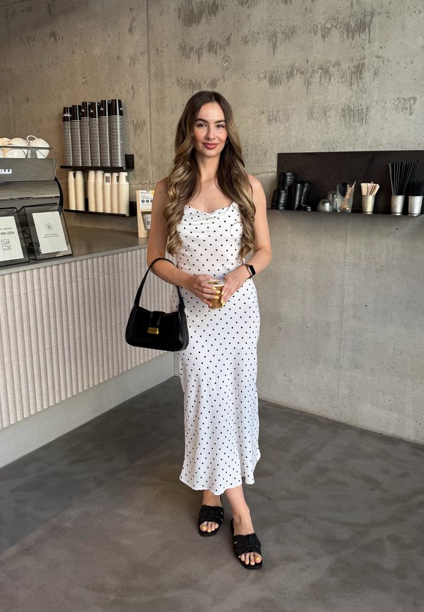 Vestido branco com bolinhas, com decote drapeado, combinado com uma bolsa preta e sandálias planas pretas. Fundo de concreto com decoração minimalista.