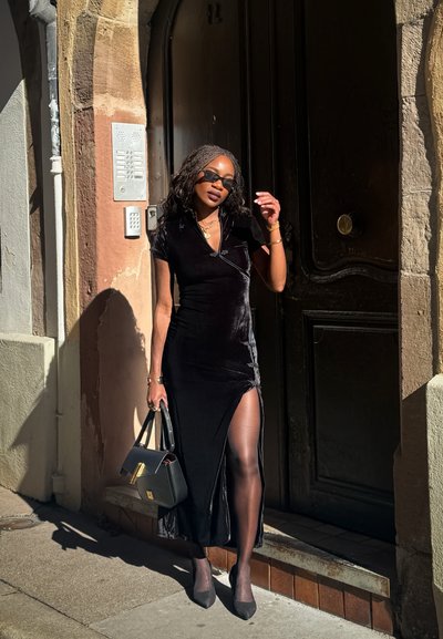 Vestido envolvente de terciopelo negro con una abertura alta, mangas cortas, combinado con un bolso negro y gafas de sol. Llevado con medias transparentes y tacones.