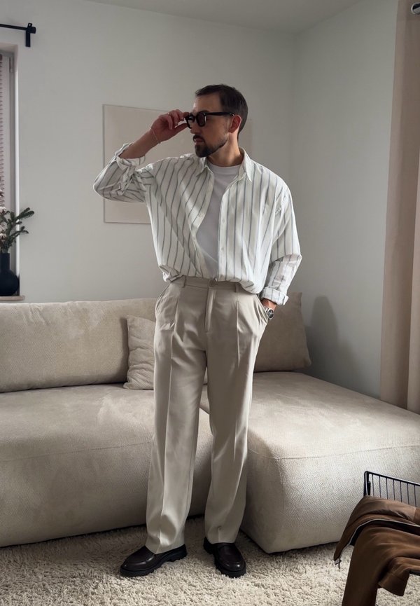 Uomo con occhiali da sole neri regolati, indossa una camicia a righe bianche, pantaloni beige e scarpe nere, in piedi in un soggiorno moderno.