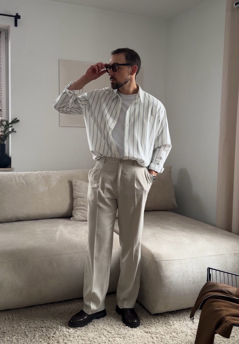 Muž nastavujúci čierne slnečné okuliare, oblečený v bielom pruhovanom tričku, béžových nohaviciach a čiernych topánkach, stojaci v moderne zariadenej obývacej izbe.