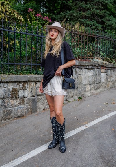 Joven mujer con un sombrero beige, camiseta negra de gran tamaño, pantalones cortos blancos de encaje, botas vaqueras negras y llevando un bolso negro junto a un muro de piedra.