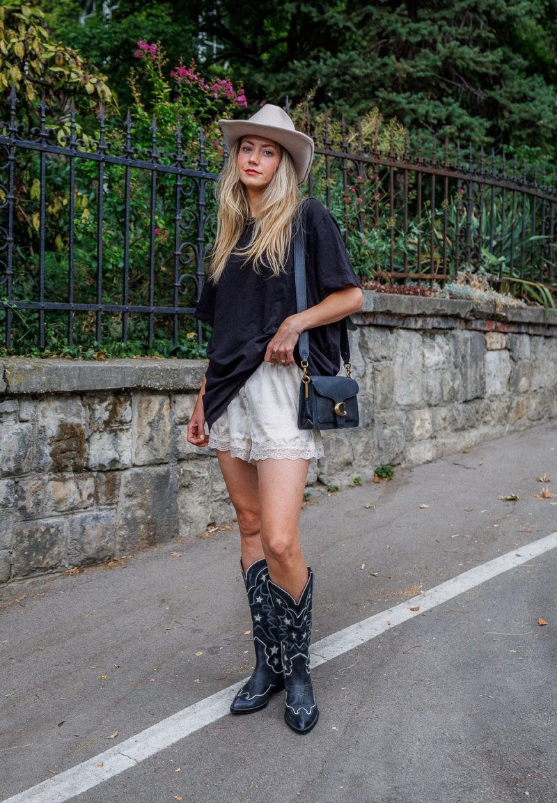 Jeune femme portant un chapeau beige, un t-shirt noir oversize, un short en dentelle blanche, des bottes de cowboy noires, et portant un sac à main noir près d'un mur en pierre.