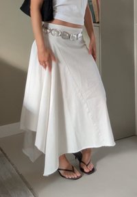 Witte asymmetrische rok van gladde stof, met een gebogen zoom en een zilveren ronde riem. Draag met zwarte flip-flops.
