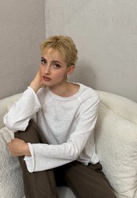 Giovane con capelli biondi corti seduto su una sedia bianca dalla superficie strutturata, indossa una camicia bianca a maniche lunghe e pantaloni marroni contro una parete strutturata.