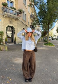 Młoda kobieta o kręconych blond włosach poprawia kaptur jasnoniebieskiej krótkiej koszuli, ubrana w brązowe szerokie spodnie, stojąca na miejskim chodniku.