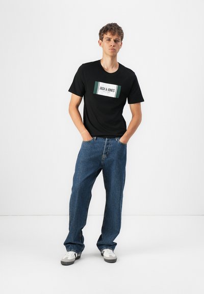 Camiseta negra de manga corta con un logo blanco y verde, combinada con pantalones vaqueros azules holgados y zapatillas blancas. Fondo blanco liso.
