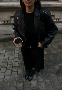 Czarna skórzana kurtka motocyklowa na czarnym topie, w komplecie z czarną spódnicą i botkami do kostki. Trzyma kubek z kawą z czarną pokrywką. Brukowana nawierzchnia.