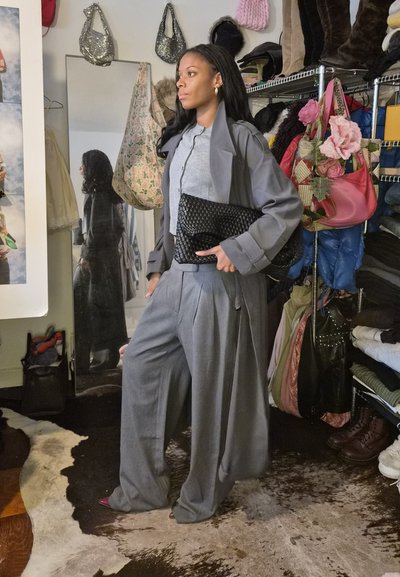 Mujer con traje gris y abrigo tipo trench, sosteniendo un bolso negro tejido, de pie en una habitación con bolsas colgadas y estantes de ropa.