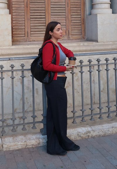 Cárdigan rojo de canalé sobre una blusa gris, combinado con pantalones de pierna ancha de cuadros negros. Sosteniendo una taza de café, con un bolso negro colgado del hombro.