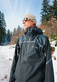 Must veekindel jakk, millel on hallid aktsendid ja tõmblukuga taskud, kõrge kraega. Kandes beeži kootud mütsi ja päikeseprille.