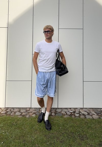 Hombre con camiseta blanca, pantalones cortos azules con rayas, zapatos negros y gafas, de pie sobre césped frente a una pared blanca de paneles sosteniendo una bolsa negra.